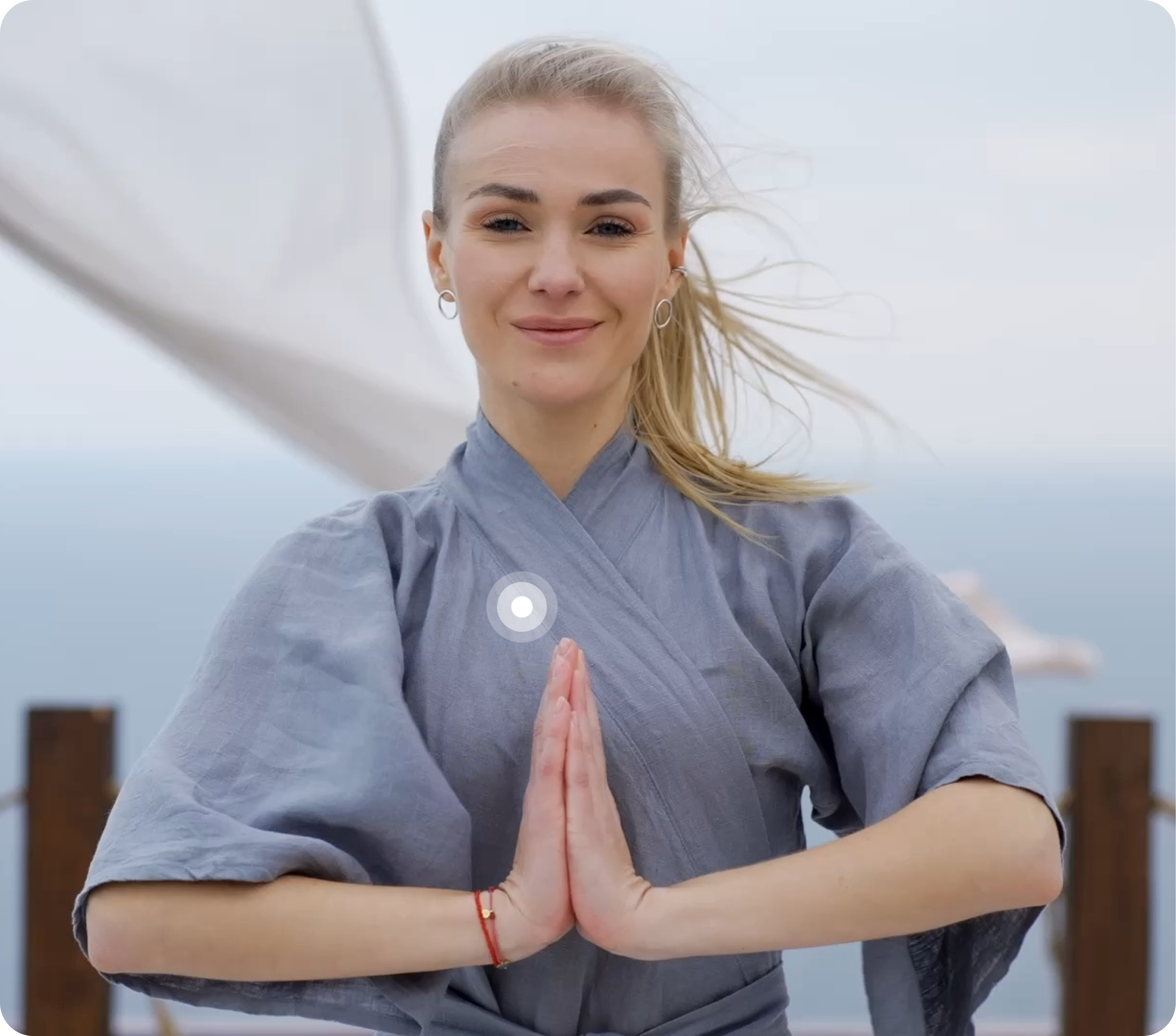 Woman with blonde hair meditating outdoors wearing a gray robe with wooden fence background